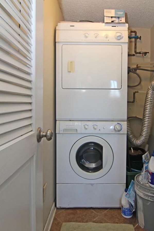 Un appareil compact, blanc, composé d’une laveuse et d’une sécheuse superposées, installé dans une petite buanderie équipée d’une porte, d’un tuyau d’évacuation et d’un tapis vert sur le sol.