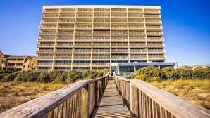 A multi-story hotel with a central courtyard, a wooden boardwalk leading to the entrance, and a clear blue sky. - Pelican Watch 907-Lovely end unit with views galore and elevators (Carolina Beach)