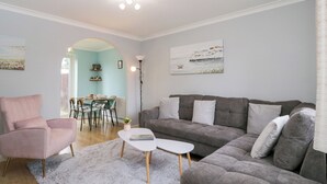 A modern living room with a grey sofa, a pink armchair, and a white coffee table. There is a dining area with a table and chairs in the background. - 20 Honeycroft (Cheltenham)