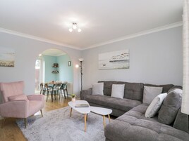 Una sala de estar moderna con un sofá gris, un sillón rosa y una mesa de centro blanca. Se observa un área de comedor con una mesa y sillas al fondo.