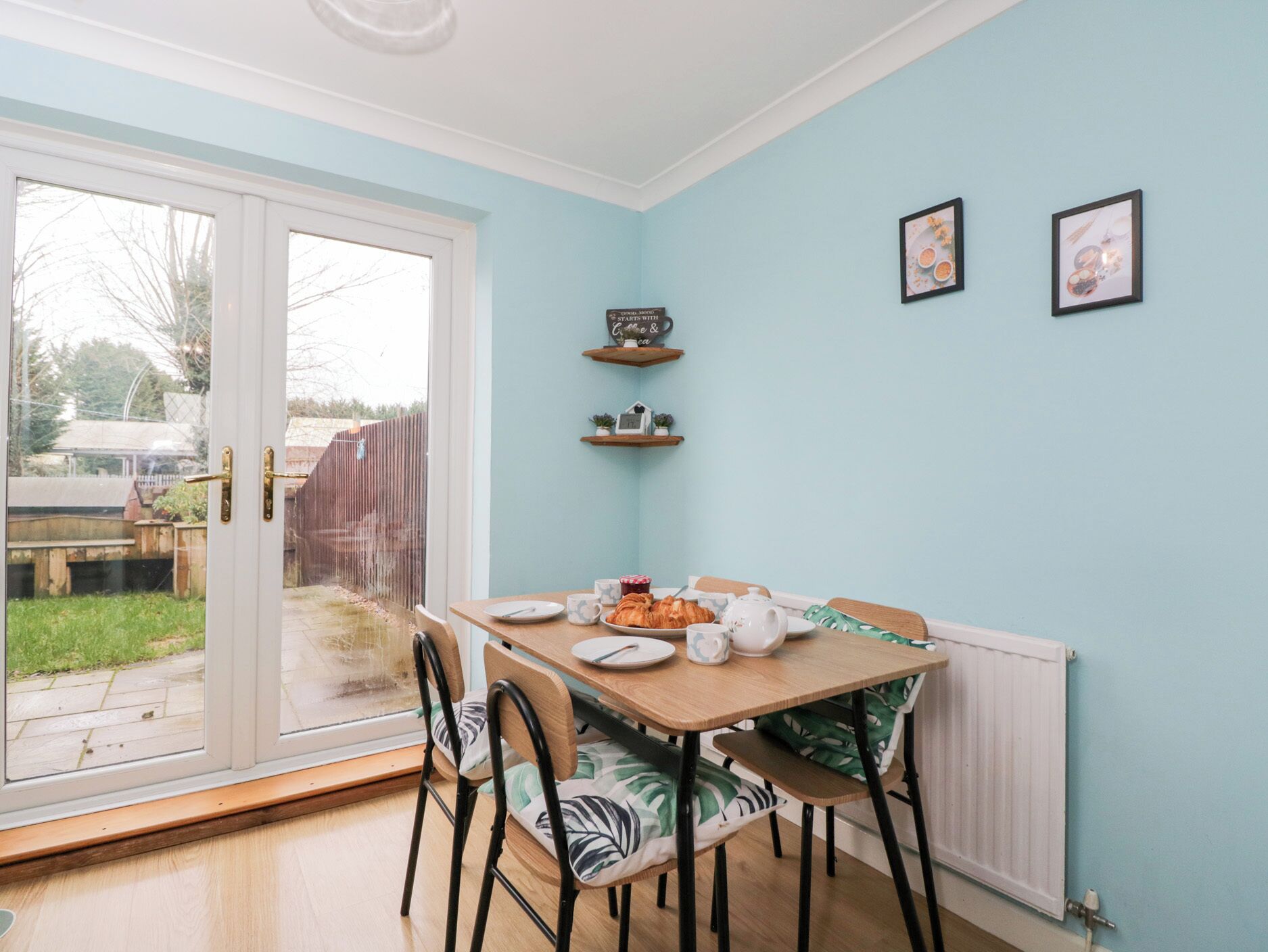 A dining area with a table set for two, chairs, a window with a view of the outside, and a wall with two framed pictures.