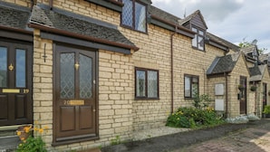 A two-story stone house with a dark brown front door, numbered 20, and a small garden with flowers and a potted plant. - 20 Honeycroft (Cheltenham)