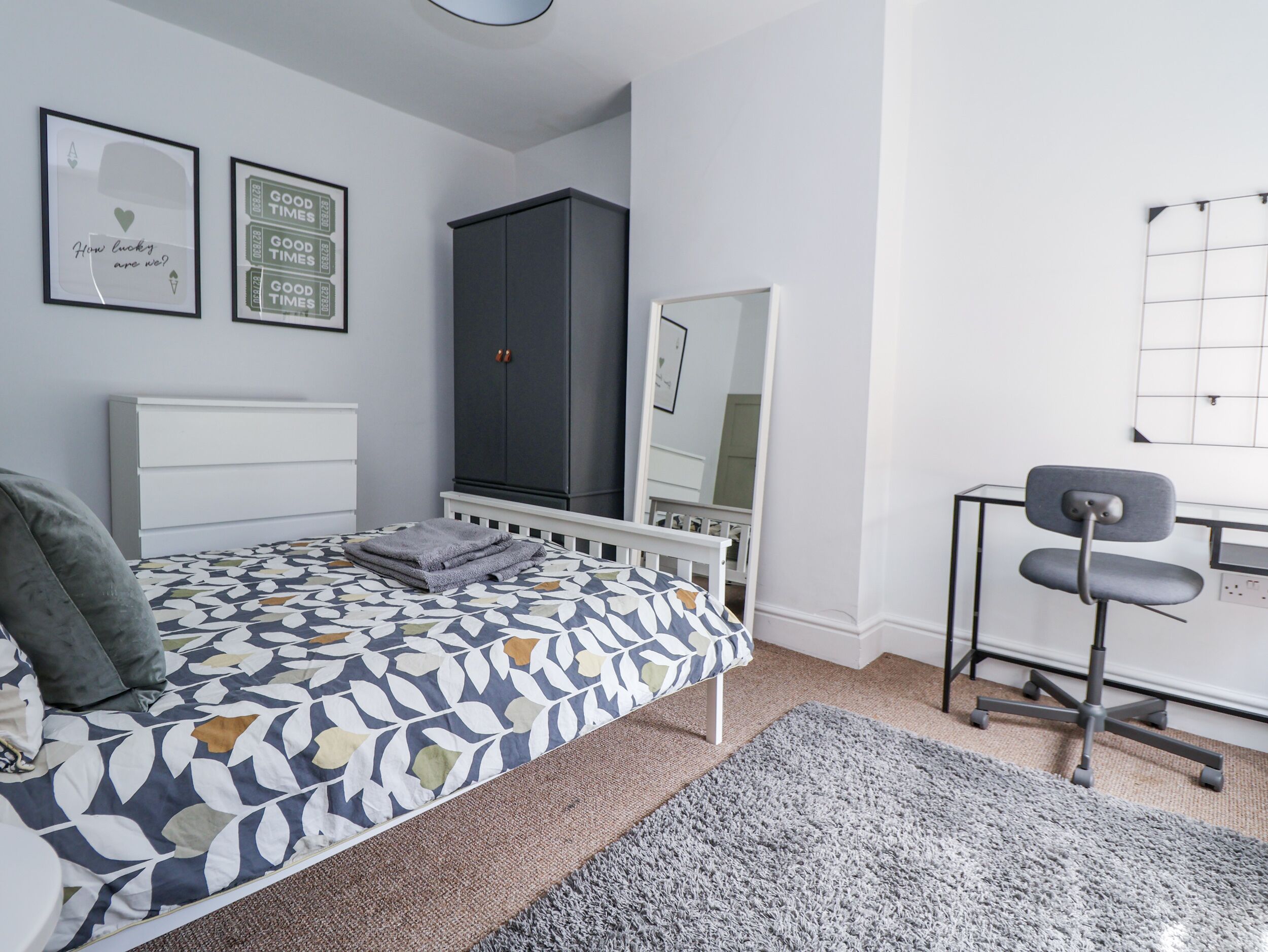 A bedroom with a bed, a grey wardrobe, a desk with a chair, and two framed motivational quotes on the wall.