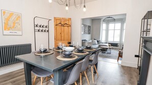 A modern dining area with a long table set for six, surrounded by grey chairs with wooden legs. A wooden door with a coat rack is visible in the background.