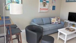 Un salon avec un canapé gris, un fauteuil en cuir noir, une table basse en bois et un téléviseur posé sur un meuble blanc. Il y a des affiches au mur, dont une sur laquelle on peut lire « FLORIDE » et une autre avec l’inscription « MANGER. SE PRÉLASSER SUR LA PLAGE. PRENDRE LE SOLEIL ».