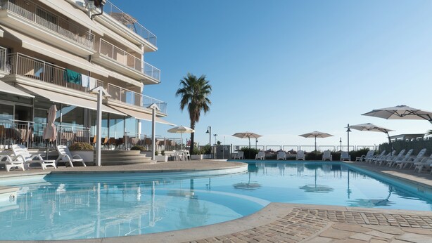 Una piscina con sillas de descanso y sombrillas, una palmera y un edificio de varios pisos con balcones.