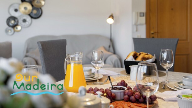 Una escena de desayuno con una jarra de jugo de naranja, copas de vino, un bol de uvas y una cesta de croissants sobre una mesa.