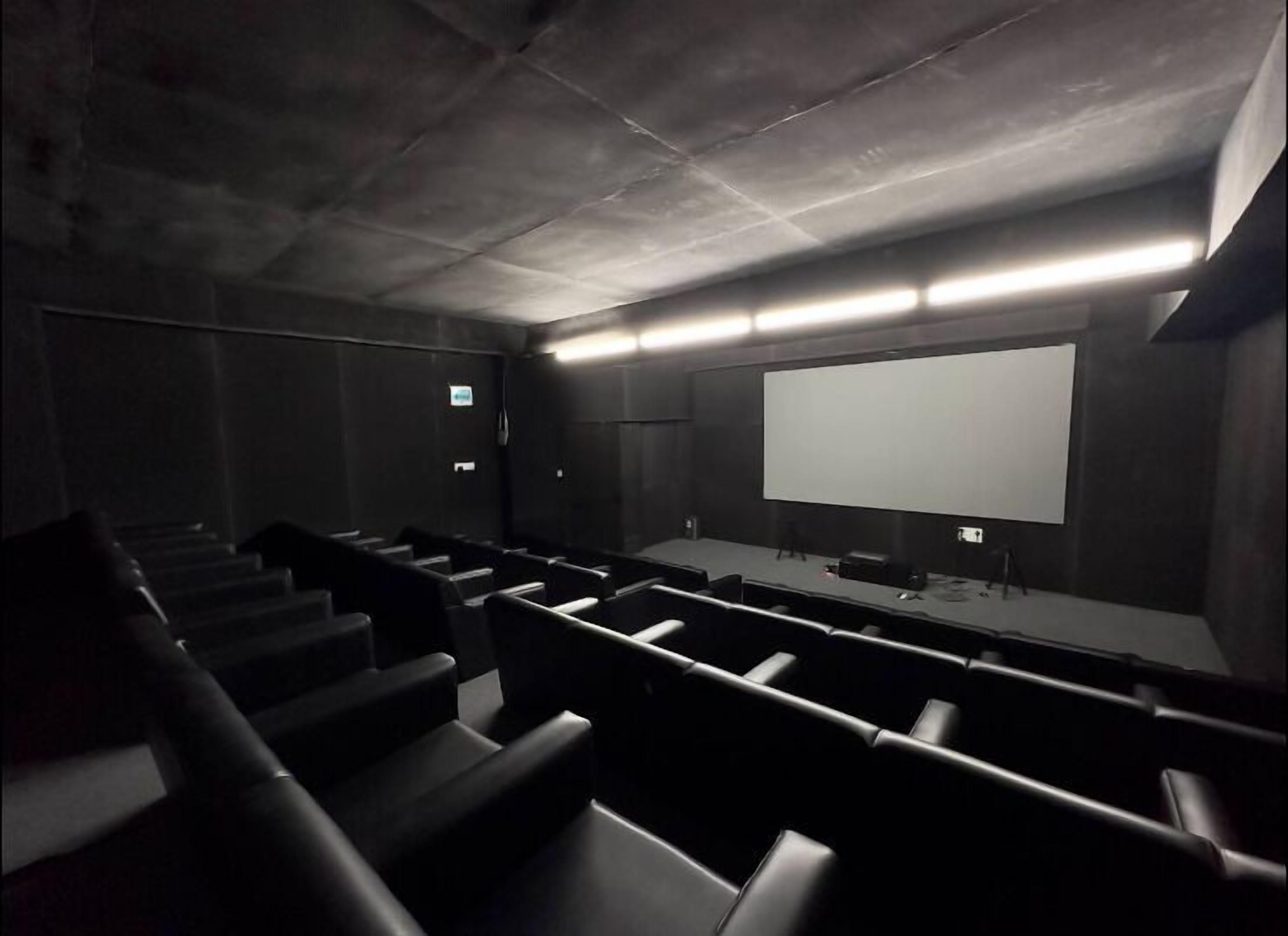 A dark, empty movie theater with rows of black seats, a large screen, and a staircase leading to the upper levels.