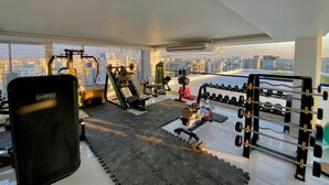 Un gimnasio con diferentes equipos de ejercicio, un ventanal con vista a la ciudad y una silla de ejercicio negra.