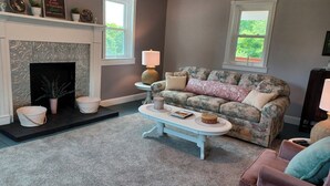 A living room with a fireplace, a floral sofa, a coffee table, and a view of greenery outside. - Heritage Homeplace in the Highlands (Farmington)