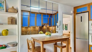A kitchen with a wooden table, chairs, a refrigerator, and a window with blue-tinted glass.