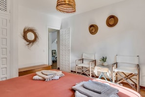 Une chambre à coucher comprenant un lit, deux chaises, une petite table, une plante en pot et une porte donnant sur une autre pièce.