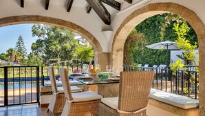 A dining area with wicker chairs and a table set for two, overlooking a swimming pool and greenery.