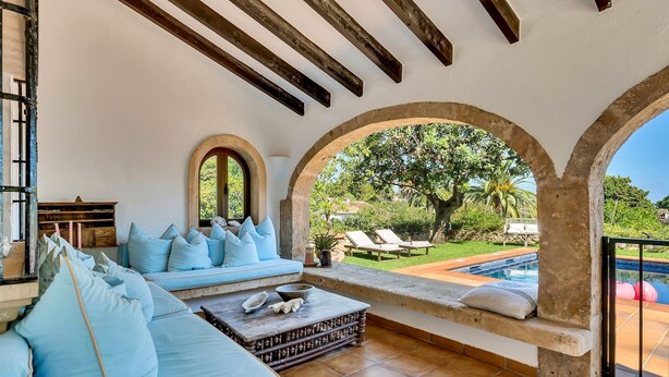 A sunlit room with a stone bench, a cushioned seating area, and a view of a swimming pool and garden.