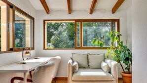 A cozy room with a desk, a cushioned seating area, large windows, and a potted plant.