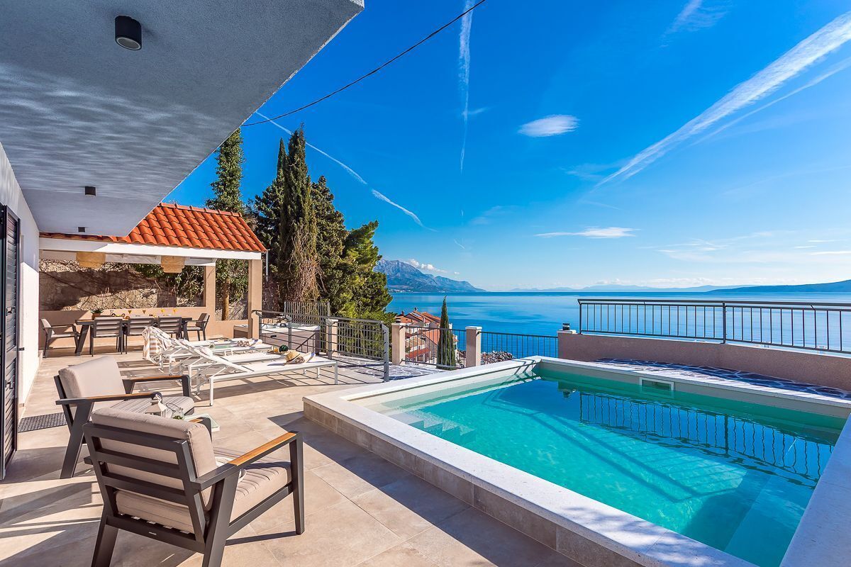 A pool area with a clear view of the sea and mountains, outdoor furniture, and a covered seating area.