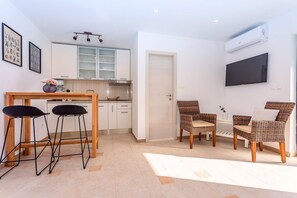 A modern kitchen with white cabinets, a wooden dining table, and two black bar stools. There is a TV mounted on the wall and a wicker chair set in the living area. - Villa Tadsi - Four Bedroom Villa, Sleeps 8 (Pisak)