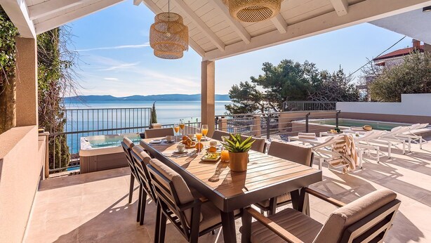 Une terrasse avec une table en bois, des chaises et un pot de fleurs. On aperçoit une piscine et des chaises longues à l’arrière-plan.