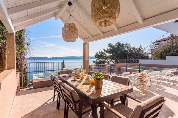 Une terrasse avec une table en bois, des chaises et un pot de fleurs. On aperçoit une piscine et des chaises longues à l’arrière-plan.