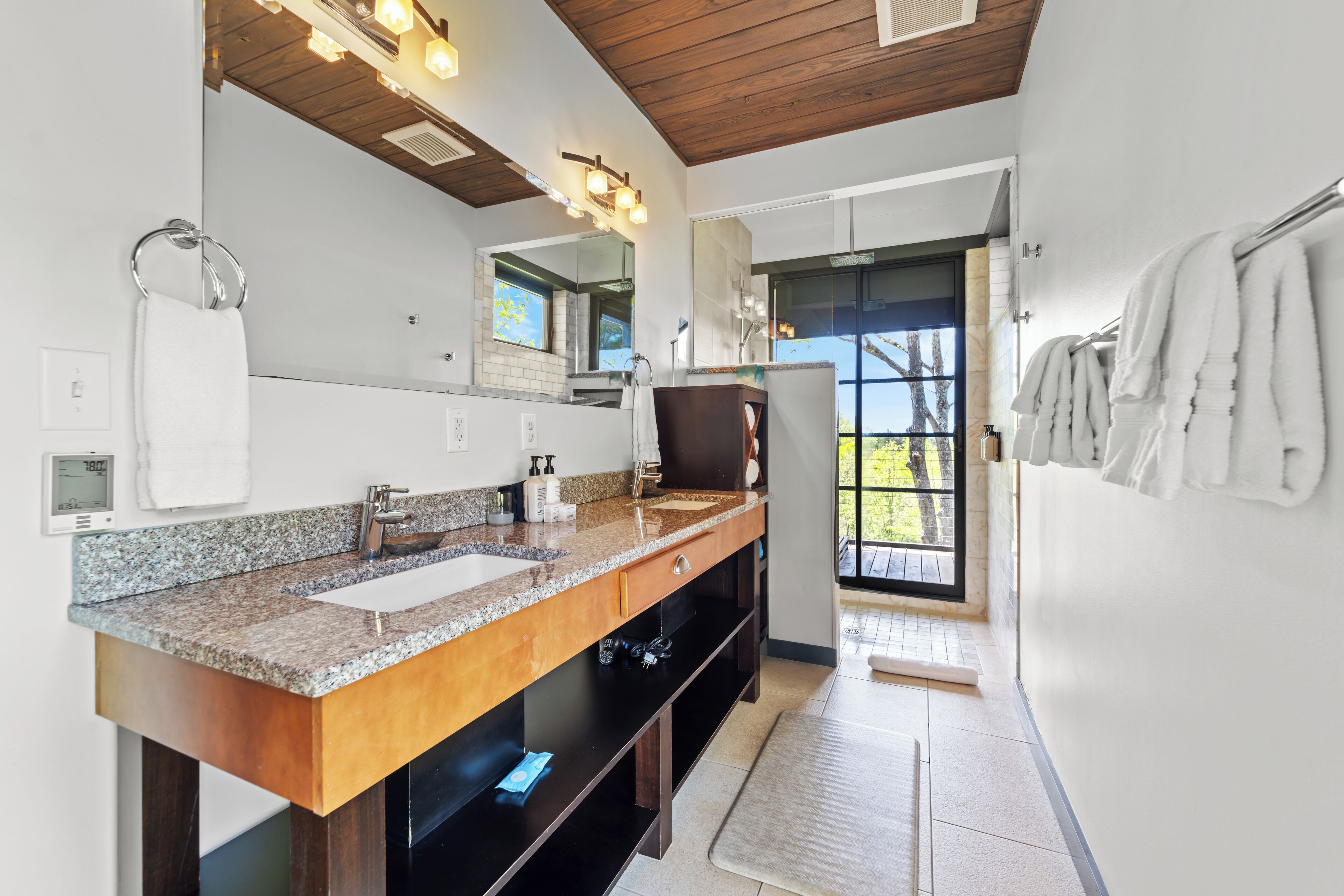 Un baño moderno con lavamanos de granito, tocador de madera, un espejo grande y vistas al exterior a través de una puerta de cristal.