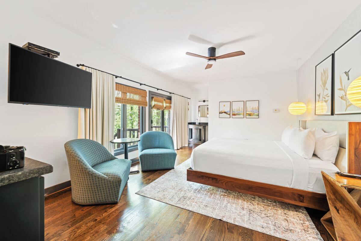 Ein modernes Hotelzimmer mit einem großen Bett, zwei Sesseln, einem Flachbildfernseher, einem Deckenventilator und einem großen Fenster mit Vorhängen.