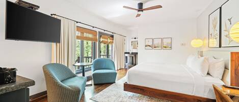 Une chambre d’hôtel moderne dotée d’un grand lit, de deux fauteuils, d’un téléviseur à écran plat, d’un ventilateur de plafond et d’une grande fenêtre avec des rideaux.