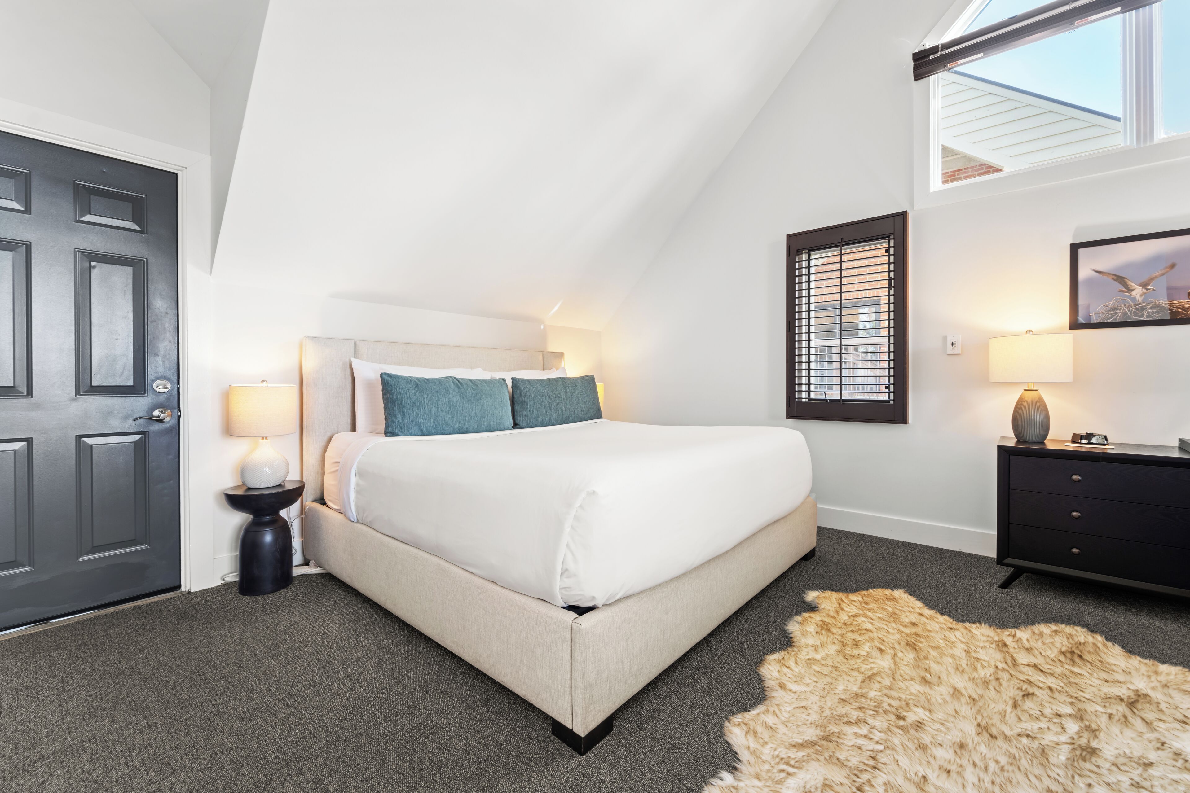 A bedroom with a bed, bedside lamps, a dark door, a window with blinds, and a rug.