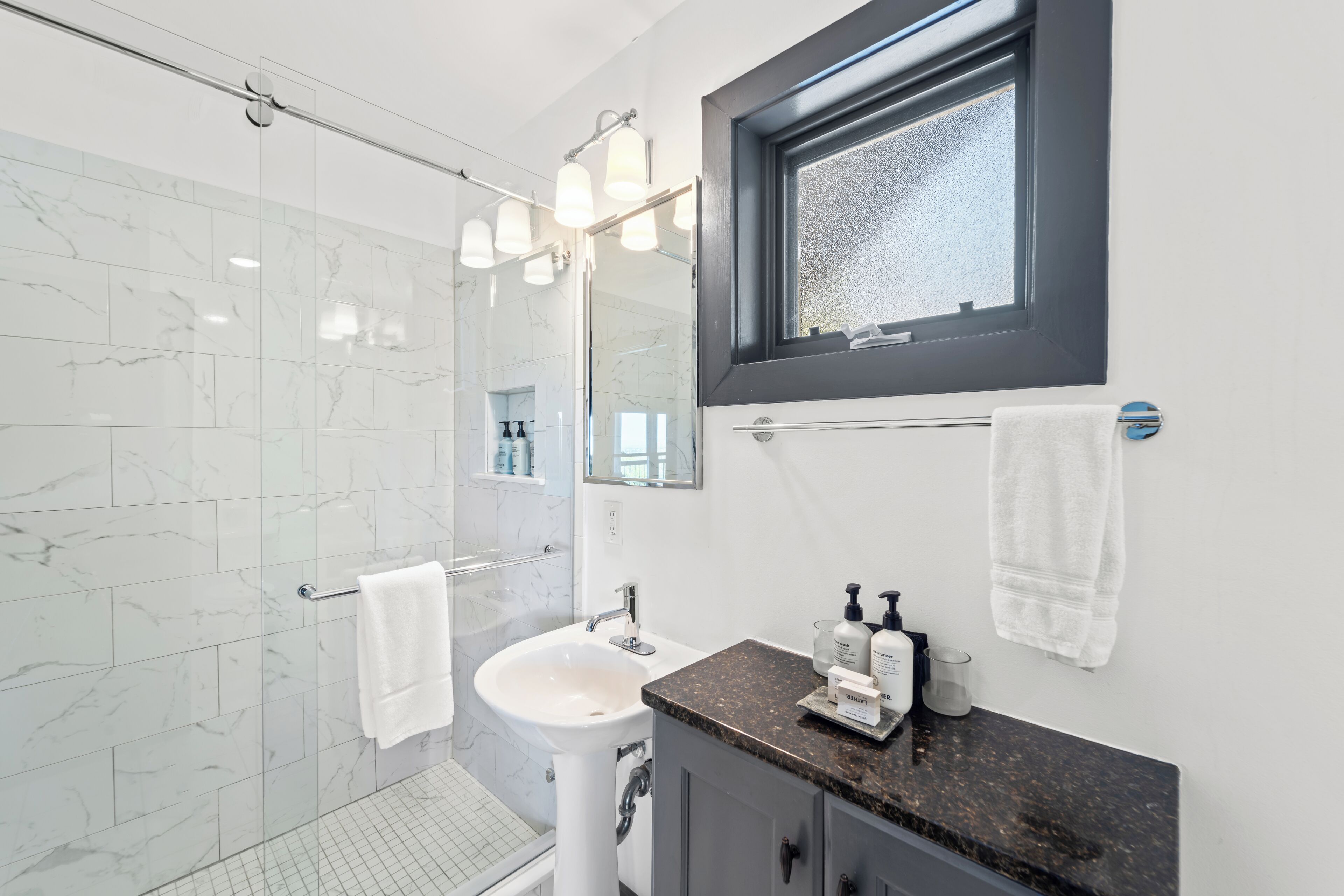 A modern bathroom with a glass shower, white sink, and dark granite countertop.