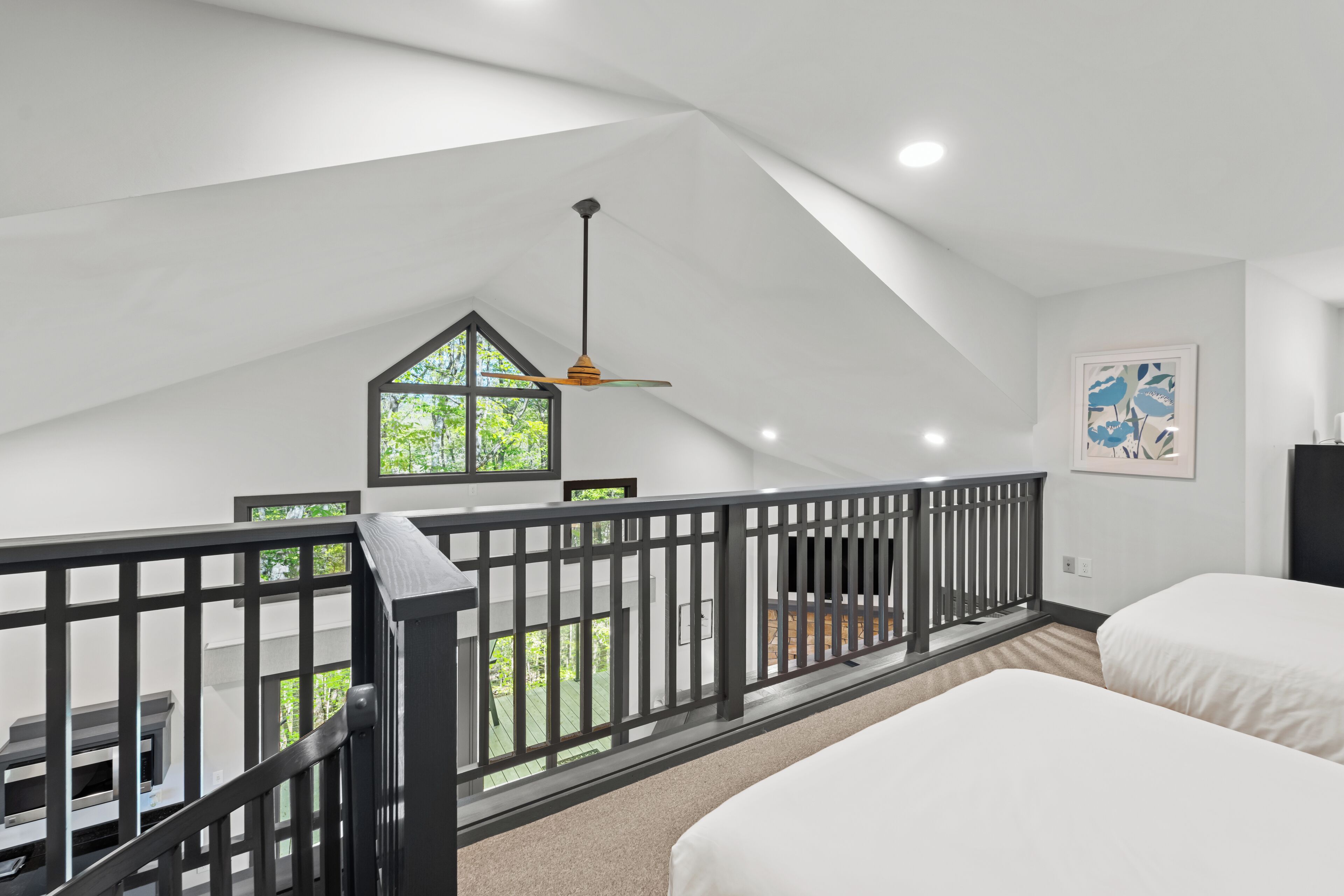 A bedroom with two beds, a balcony with a railing, a ceiling fan, and a window with a view of greenery.