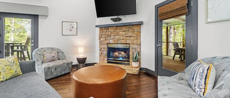 A living room with a stone fireplace, a flat-screen TV mounted above it, and a large window providing a view of the outdoors.