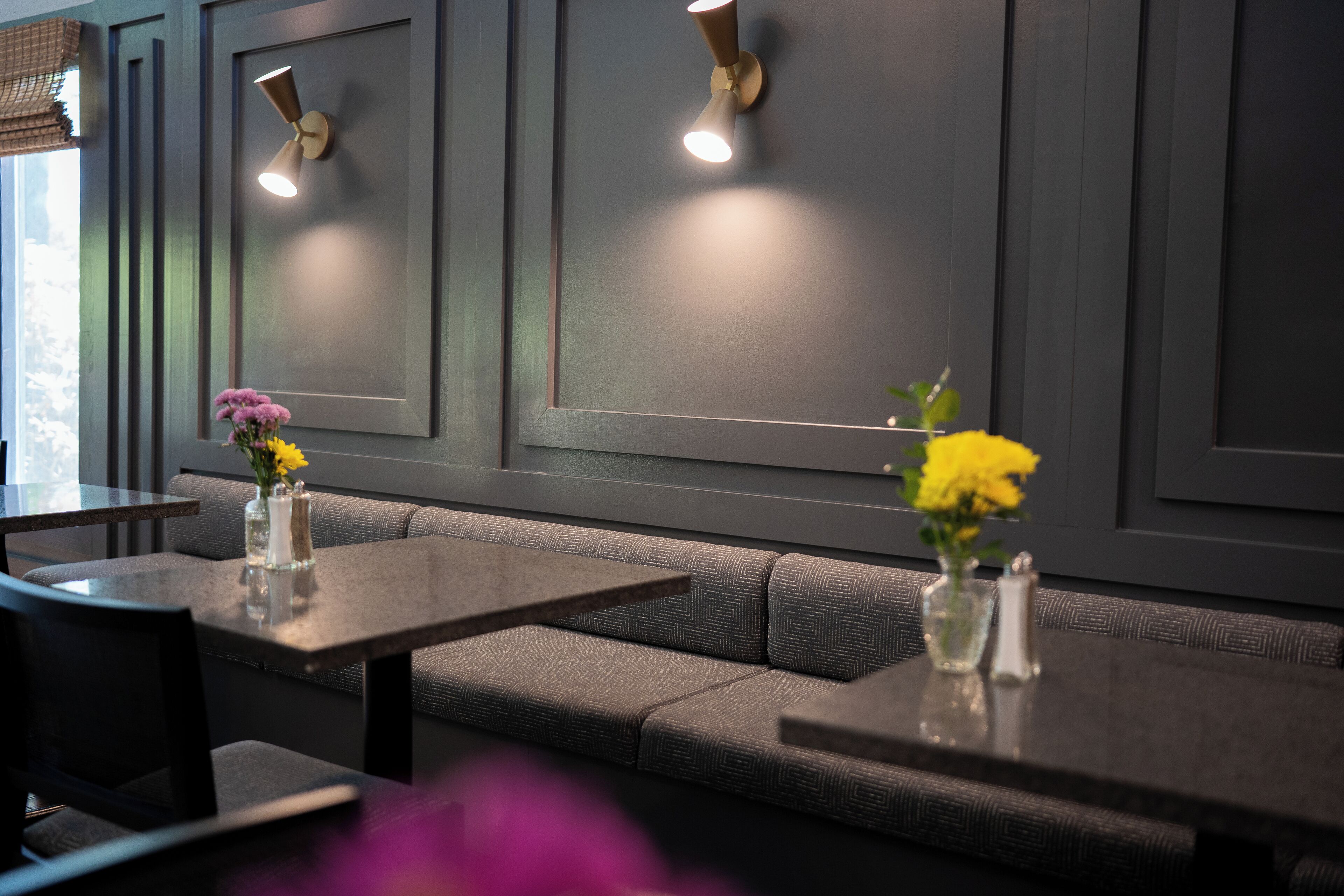 Ein modernes Interieur mit einer grauen Polsterbank und Tischen, zwei Wandleuchten sowie Vasen mit Blumen.