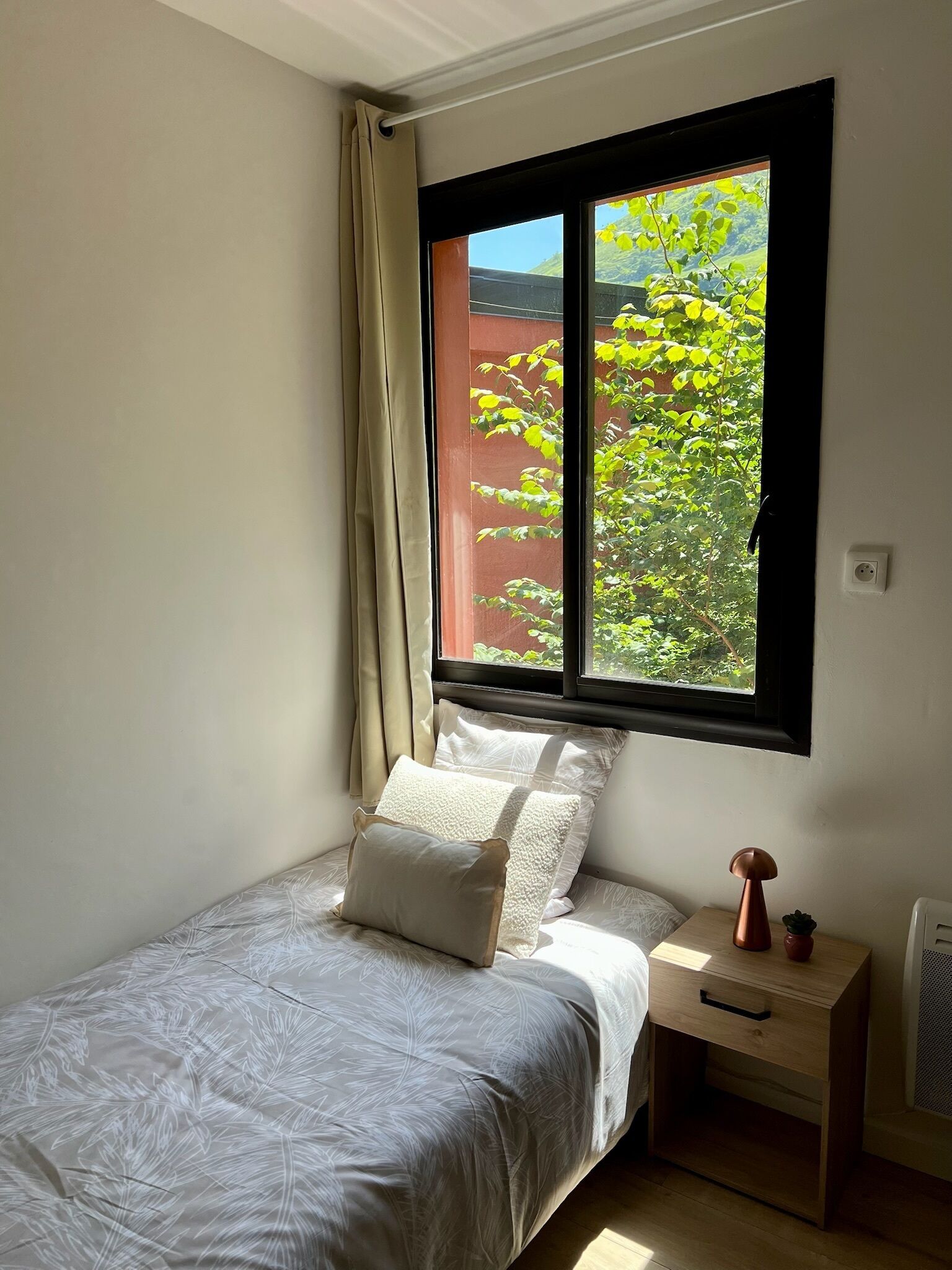 A bedroom with a bed, a nightstand, a window with a view of greenery, and a wall socket.