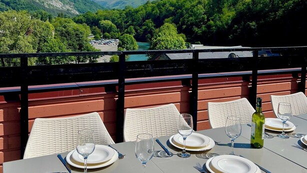 Una mesa de comedor dispuesta con platos, cubiertos y copas de vino, con vistas a un paisaje montañoso.