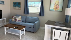 Un salon avec un canapé gris, une table basse en bois, une petite table d’appoint blanche et une petite table blanche avec une lampe. Deux tableaux ornent les murs et une fenêtre est agrémentée de rideaux bleus.