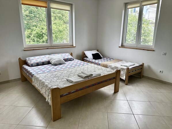 Une chambre avec deux lits en bois, chacun agrémenté de draps et d’oreillers à motifs. Deux fenêtres laissent entrer la lumière naturelle, et la chambre est dotée d’un sol carrelé.