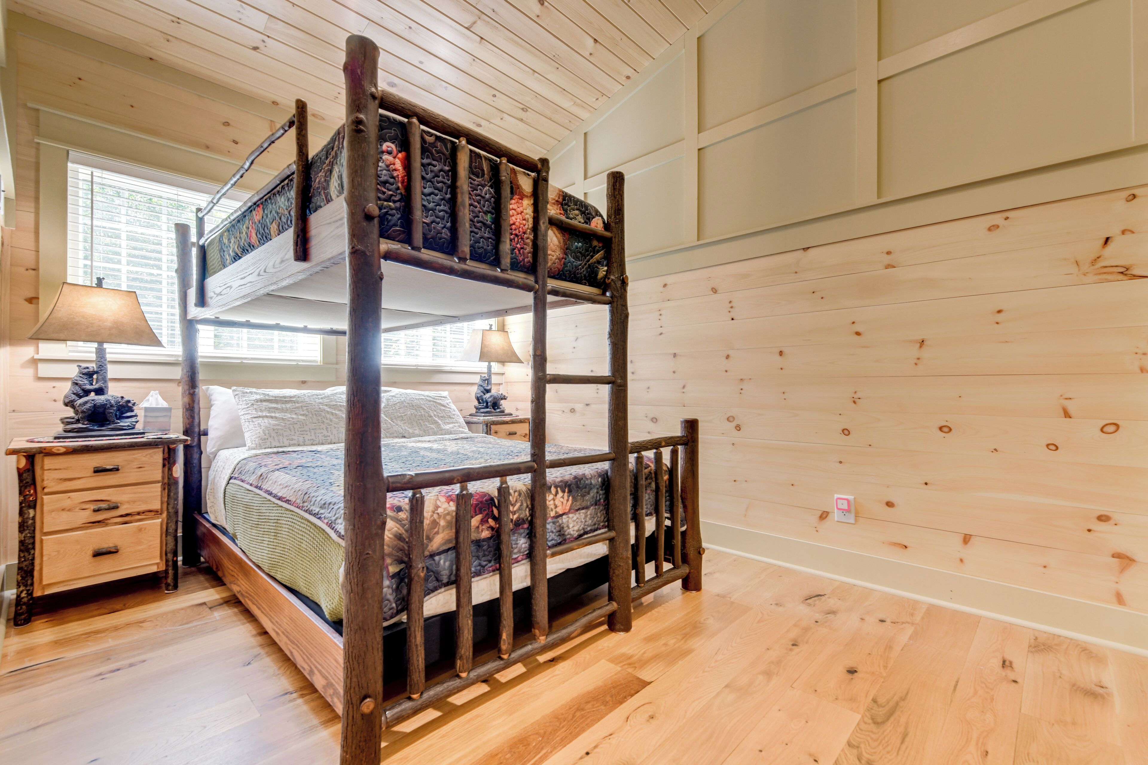 A bunk bed with a wooden frame and a patterned mattress, a bedside table with a lamp, and a window with blinds.