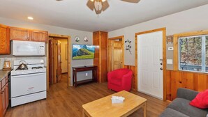 Un espacio habitable compacto con suelo de madera, una kitchenette con estufa y microondas, un televisor, una silla roja, un sofá gris y un ventilador de techo.