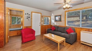 Una sala de estar acogedora, con suelo de madera, un sillón rojo, un sofá gris con cojines rojos, una mesa de centro de madera y un ventilador de techo.