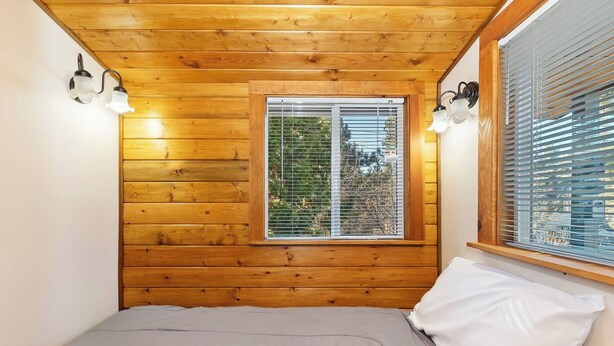 Un dormitorio con paredes de madera, una ventana con persianas, una cama con colchón gris y almohada blanca, y lámparas fijadas a la pared.