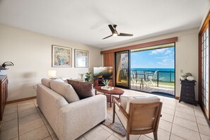 A living room with a sofa, a chair, a table, a ceiling fan, and a view of the ocean through large sliding doors.