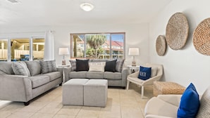 A modern living room with grey sofas, a coffee table, and a view of a residential area through large windows. - Regency Cabanas D3 (Pensacola Beach)