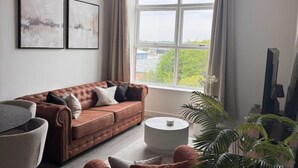 A living room with a brown leather sofa, a round coffee table, and a large window with a view of greenery and buildings.