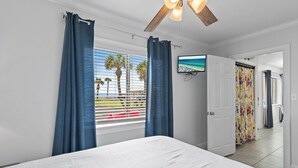 A bedroom with a bed, a ceiling fan, a TV mounted on the wall, and a view of palm trees through the window.