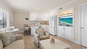 Een moderne keuken met witte kasten, een eethoek met een witte tafel en stoelen, een gezellige zithoek met een bank en fauteuil, en een groot raam met uitzicht op het strand.