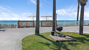 Een verhard voetpad met een barbecue en parasols, palmbomen en uitzicht op de oceaan.