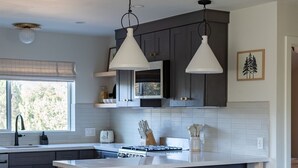 A modern kitchen with dark cabinets, white countertops, and pendant lighting. There is a dining area with a white table and a brown leather sofa with patterned cushions.