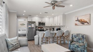 A modern kitchen with white cabinets, a microwave, and a ceiling fan. There is a dining area with a table and chairs, and a chair with a tropical pattern. - 🌴BEACH FRONT- Chair Service-Capri by the Gulf 122 (Destin)