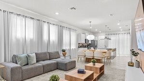 A modern living room with a grey sectional sofa, wooden coffee tables, and a dining area with a white table and chairs.