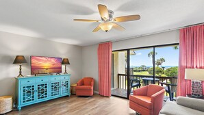 A room with a wooden floor, a ceiling fan, a large window with a view of palm trees, and a blue cabinet with a painting.
