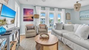 A spacious living room with a white sofa, two armchairs, a coffee table, and a ceiling fan. There are paintings on the walls, a window with curtains, and a view of the outside.